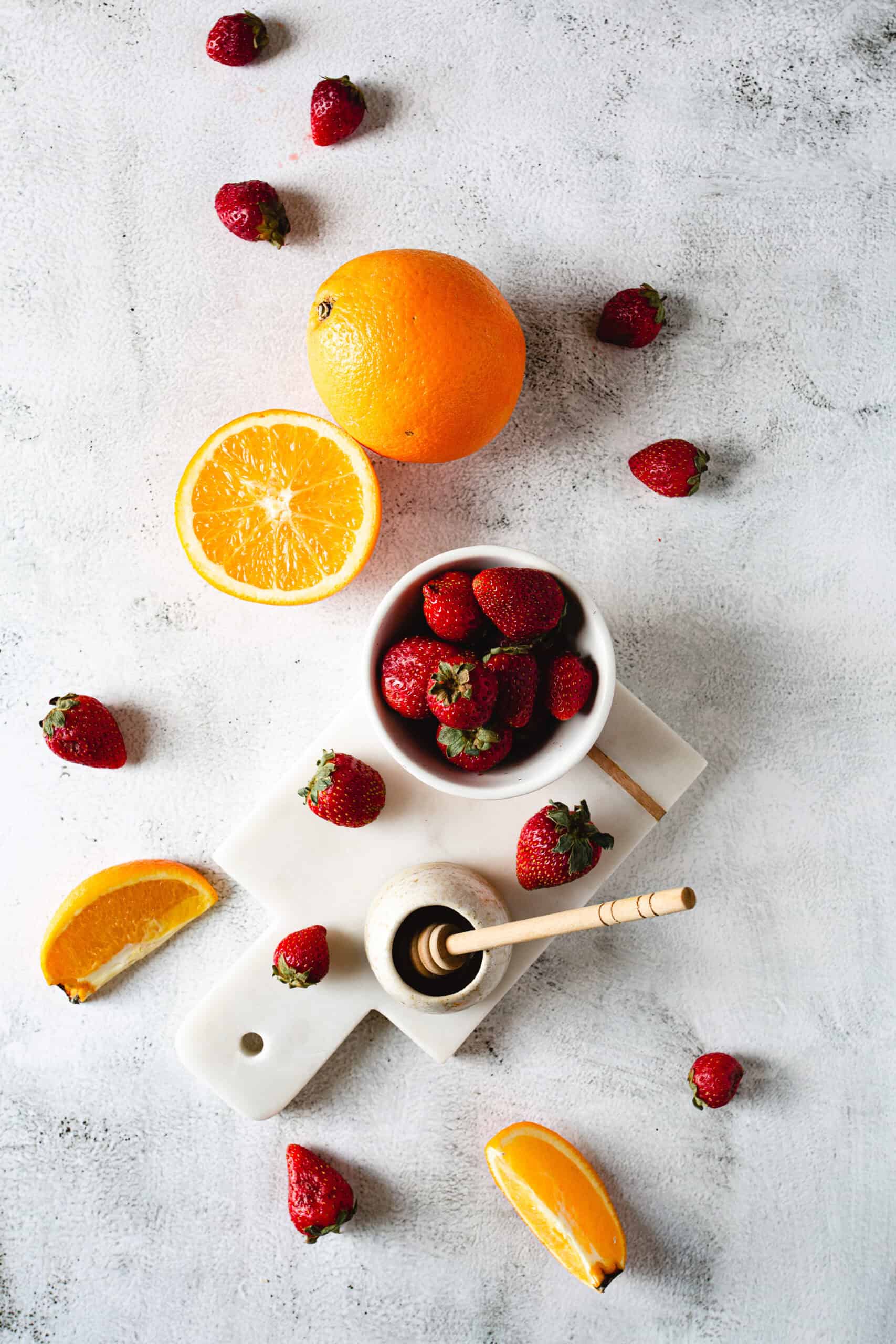 A sliced orange and a whole orange are placed on a light surface along with scattered strawberries. A bowl filled with strawberries and a honey jar with a dipper rest on a white cutting board. Orange wedges are positioned nearby.