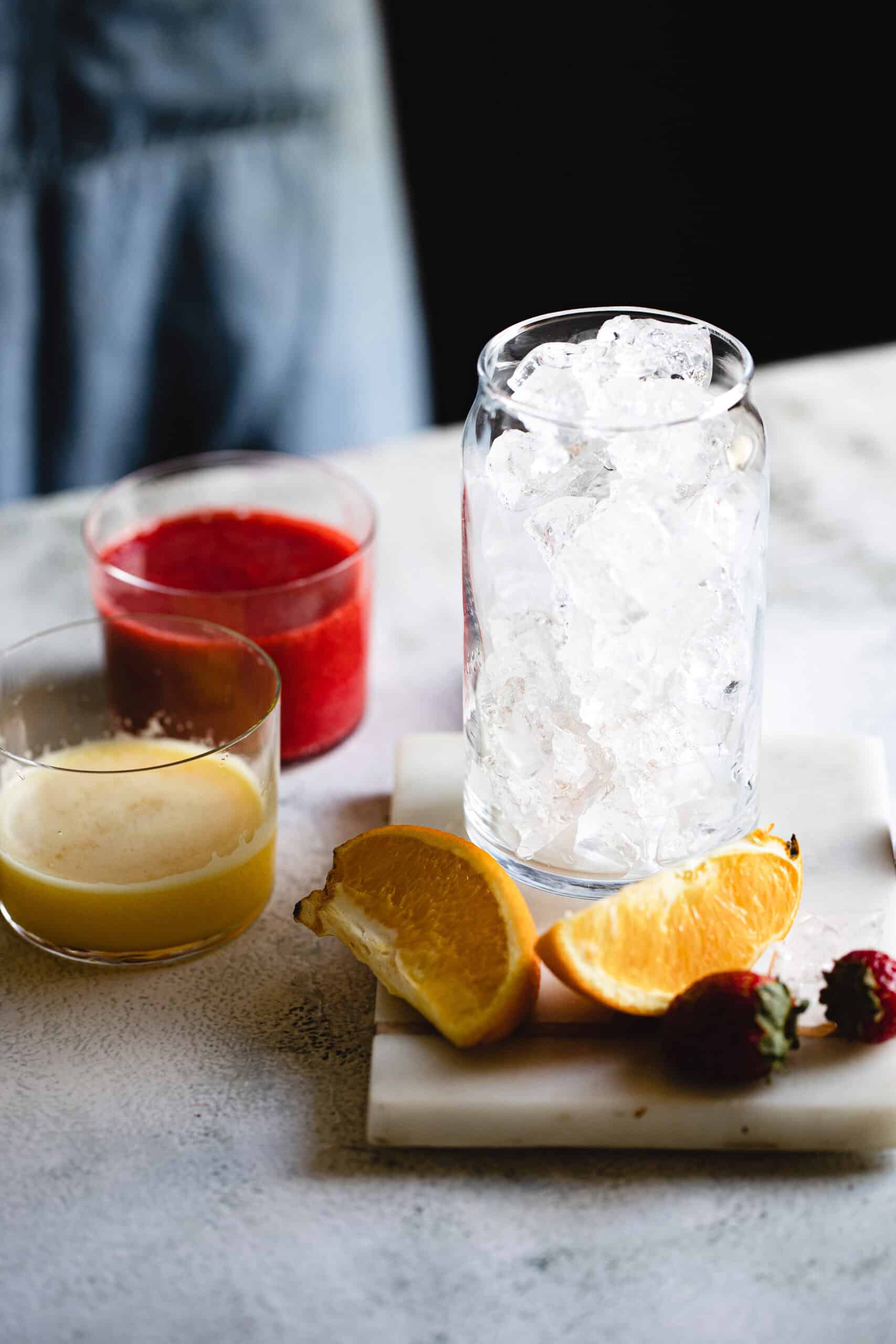 A clear glass filled with ice is on a table. Nearby are two cut orange slices, two strawberries, a small glass with red liquid, and another with yellow liquid. The background is a blurred dark gray with a hint of blue fabric.