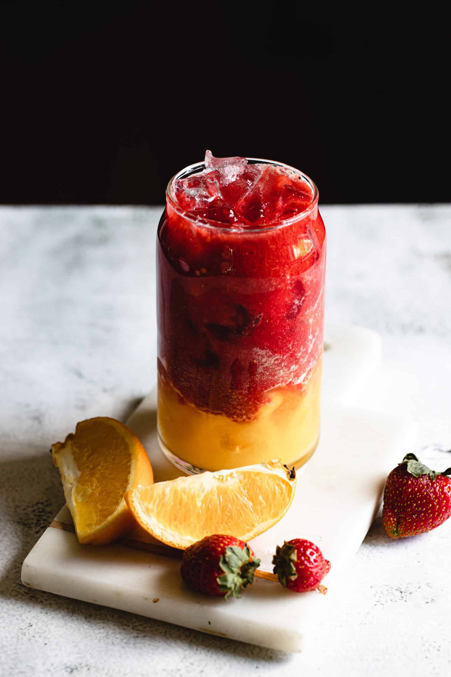 A glass filled with a layered drink, featuring a red top portion and a yellow bottom portion, is set on a white marble surface. Surrounding the glass are two halved orange wedges and two whole strawberries.