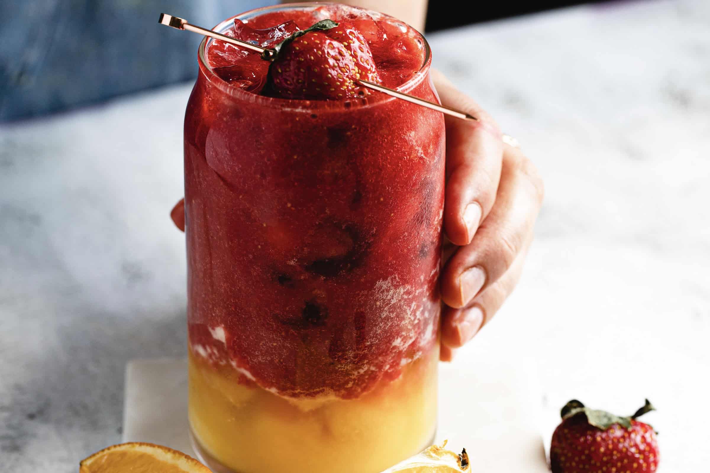 Close-up of a layered drink in a glass. The top layer is red, with strawberries and a skewer, while the bottom layer is orange. A hand holds the glass, and a lemon wedge lies on the surface nearby.