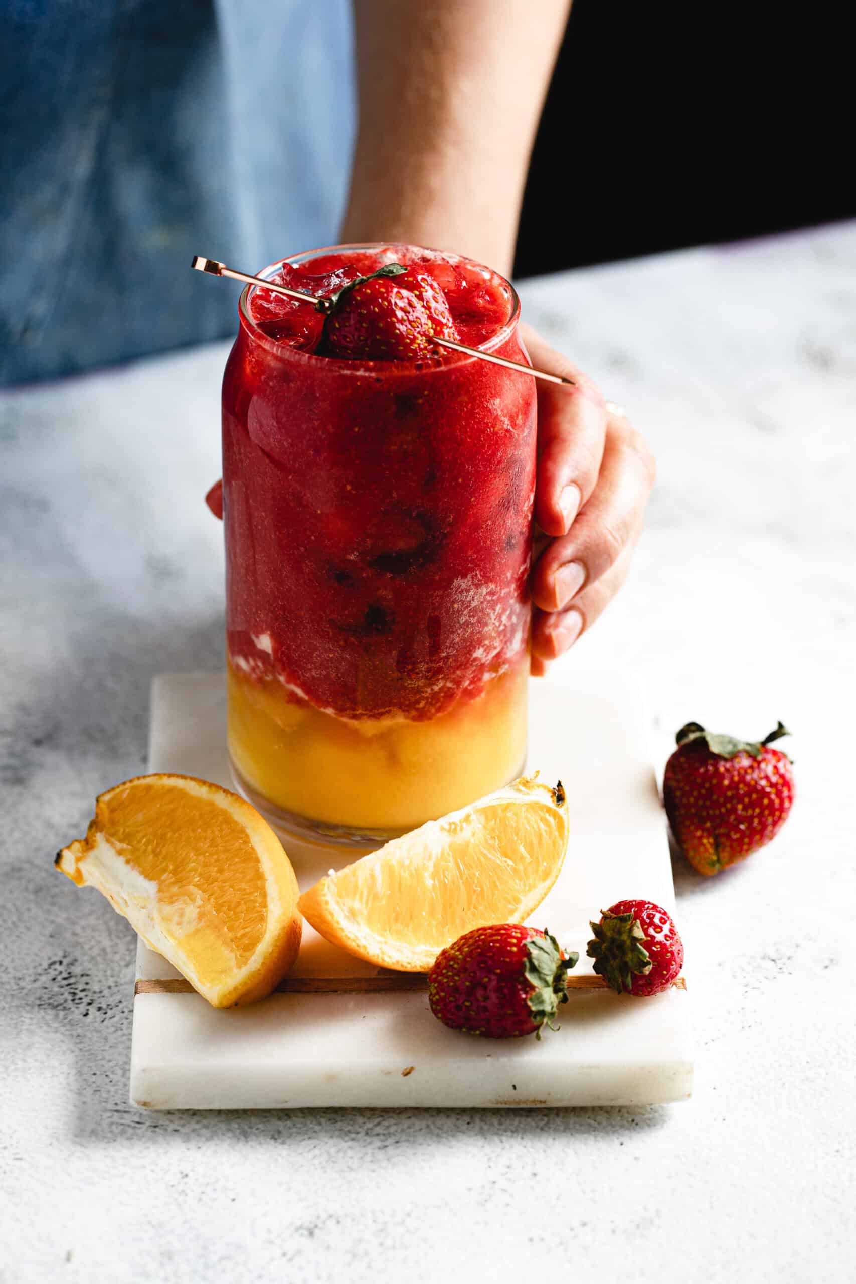 A person holds a glass filled with red and yellow layered slush, garnished with a whole strawberry and a metal straw. The glass is on a white marble board with strawberry and orange slices beside it.