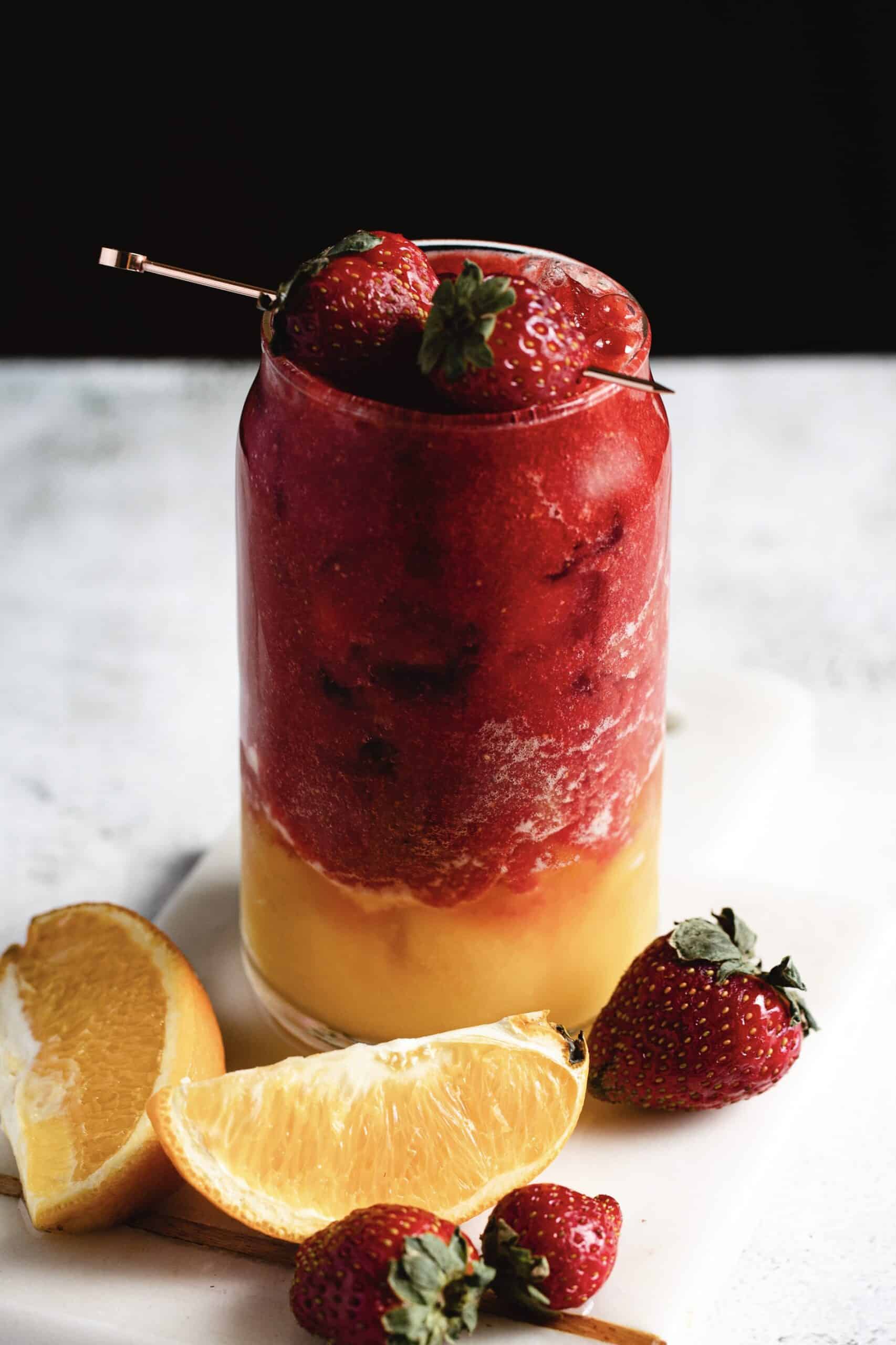 A layered smoothie in a glass, featuring a red strawberry blend on top and a yellow mango blend at the bottom. The drink is garnished with whole strawberries. Surrounding the glass are fresh strawberries and orange slices on a white surface.