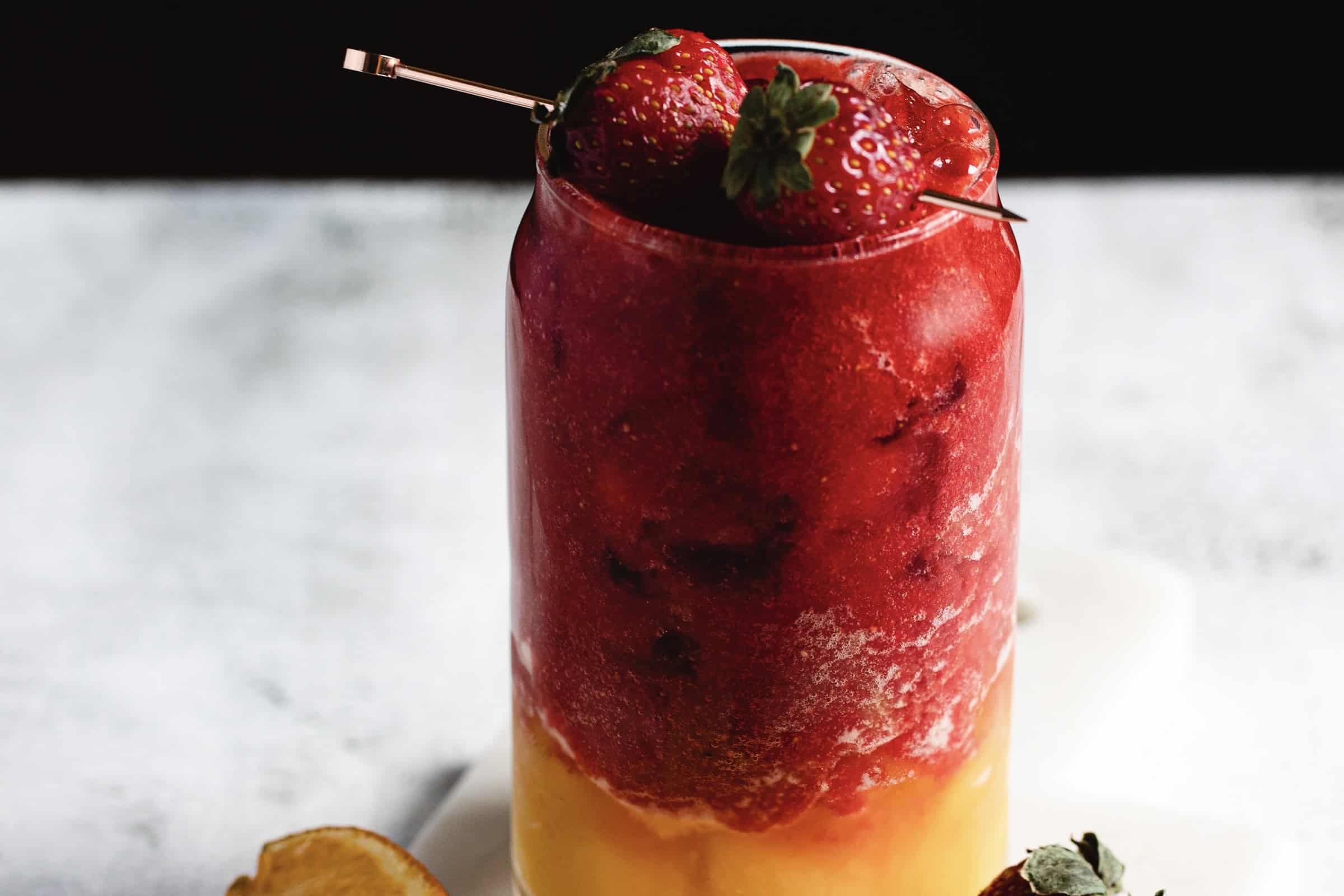 A layered drink in a glass features a vibrant red frozen mix on top of a yellow base. Two whole strawberries garnish the top, skewered with a metal pick. The glass is set against a blurred background.