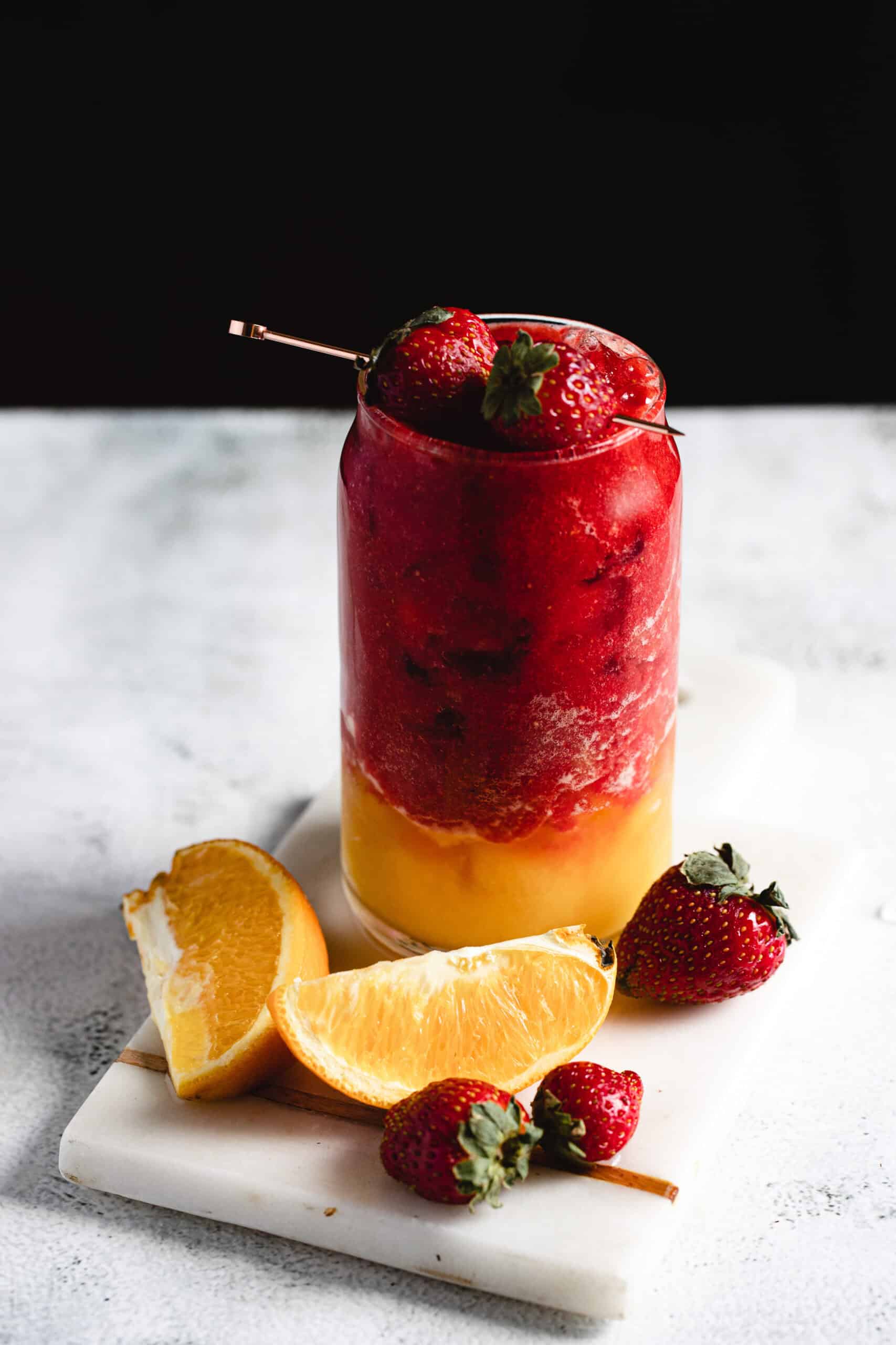 A layered drink in a glass is topped with whole strawberries. The bottom layer is orange, while the top is a deep red. The glass is on a marble surface, surrounded by sliced oranges and whole strawberries. A metal straw is inserted.