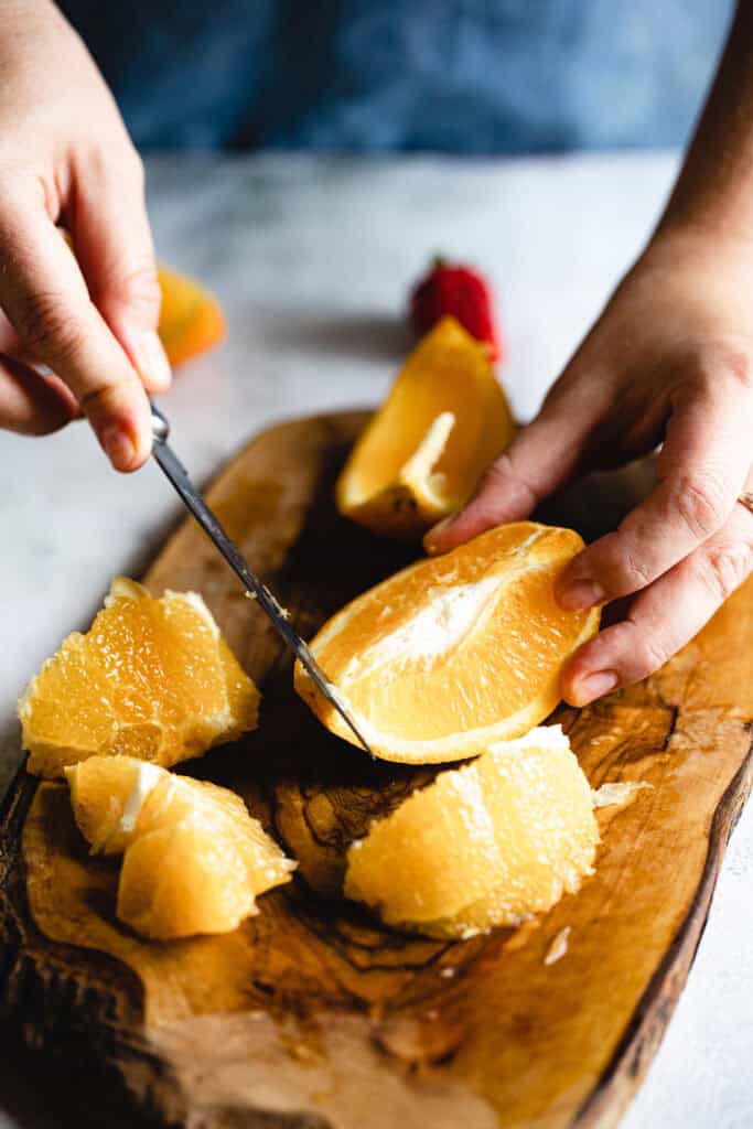 A pair of hands is slicing an orange on a wooden cutting board. Several orange segments are already cut and placed on the board. The background is blurred, with a hint of a strawberry visible. The surface is light-colored.