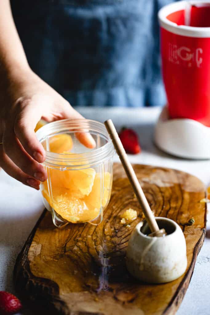 A person holds a blender cup filled with peeled orange segments over a wooden cutting board. A small marble mortar and pestle with a wooden handle is nearby. A red blender base and a strawberry are in the background.