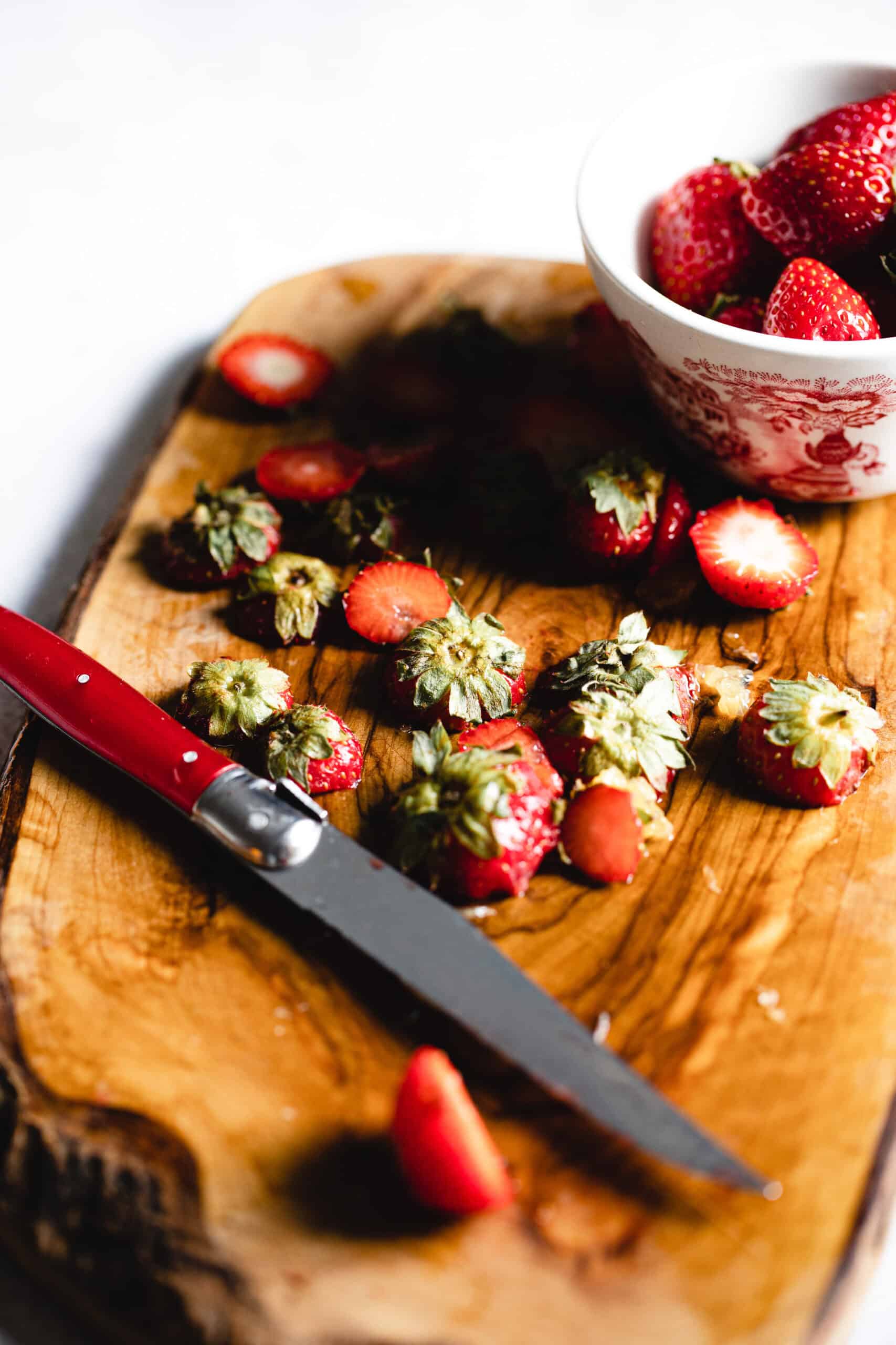 Sliced strawberries and a red-handled knife are on a wooden cutting board. A white bowl filled with whole strawberries sits nearby.