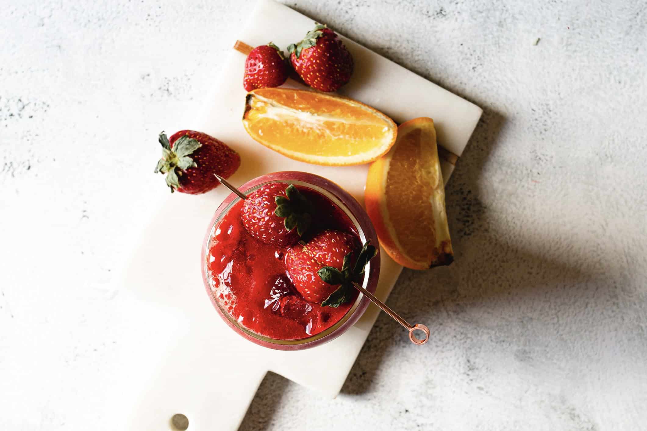 A glass filled with a red smoothie is topped with strawberries and a metal straw. It sits on a white marble board. Next to it are several whole strawberries and slices of orange on a light textured surface.