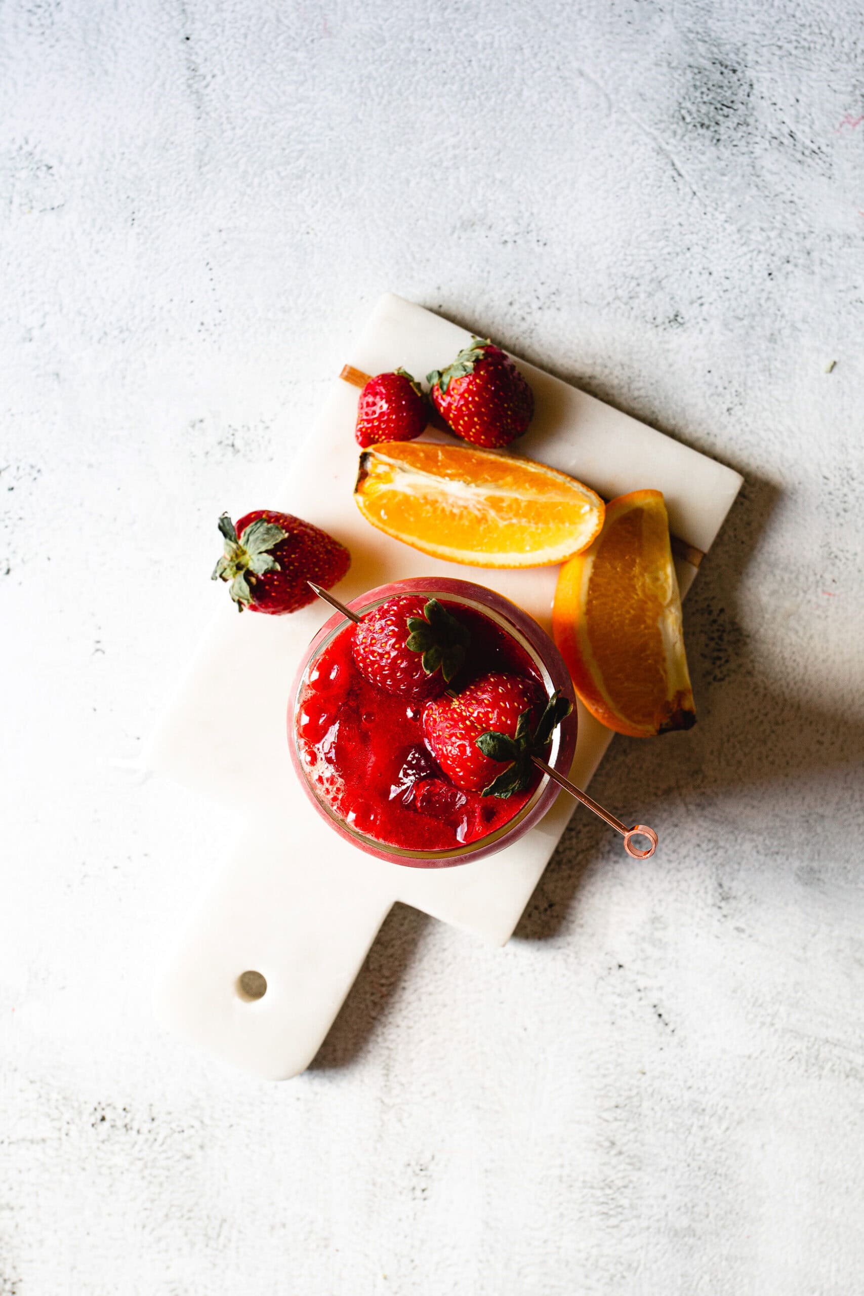 A small white cutting board on a textured surface holds a bowl of red sauce, strawberries, and orange wedges. The sauce-filled bowl and the fruits are neatly arranged on the left half of the board.