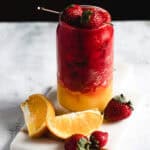 A layered drink in a glass, with red strawberry slush on top and orange slush at the bottom. Garnished with whole strawberries on top and skewered. Orange slices and strawberries are placed around the glass on a white surface.