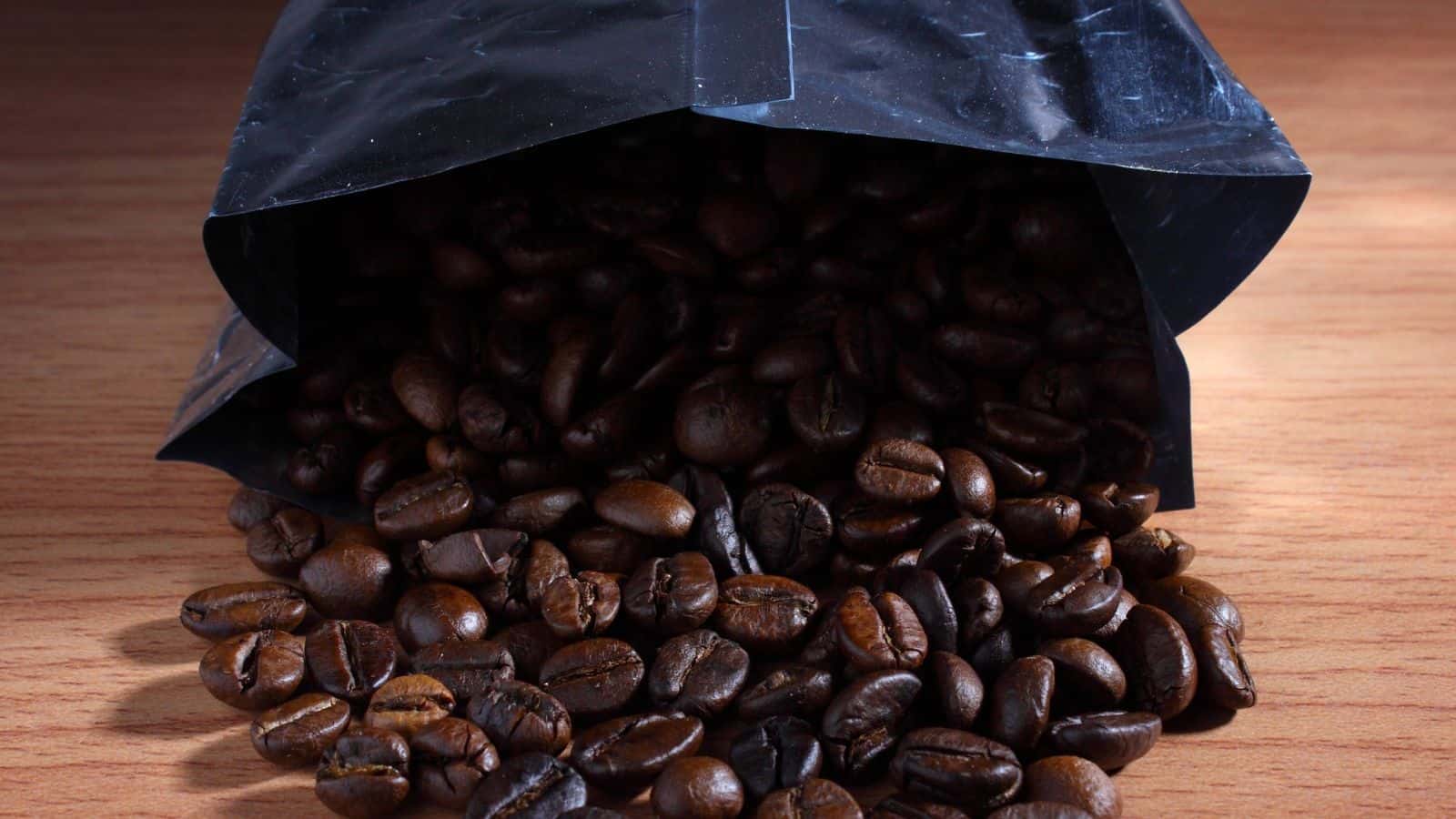 A black, open pouch lies on a wooden surface with roasted coffee beans spilling out of it.