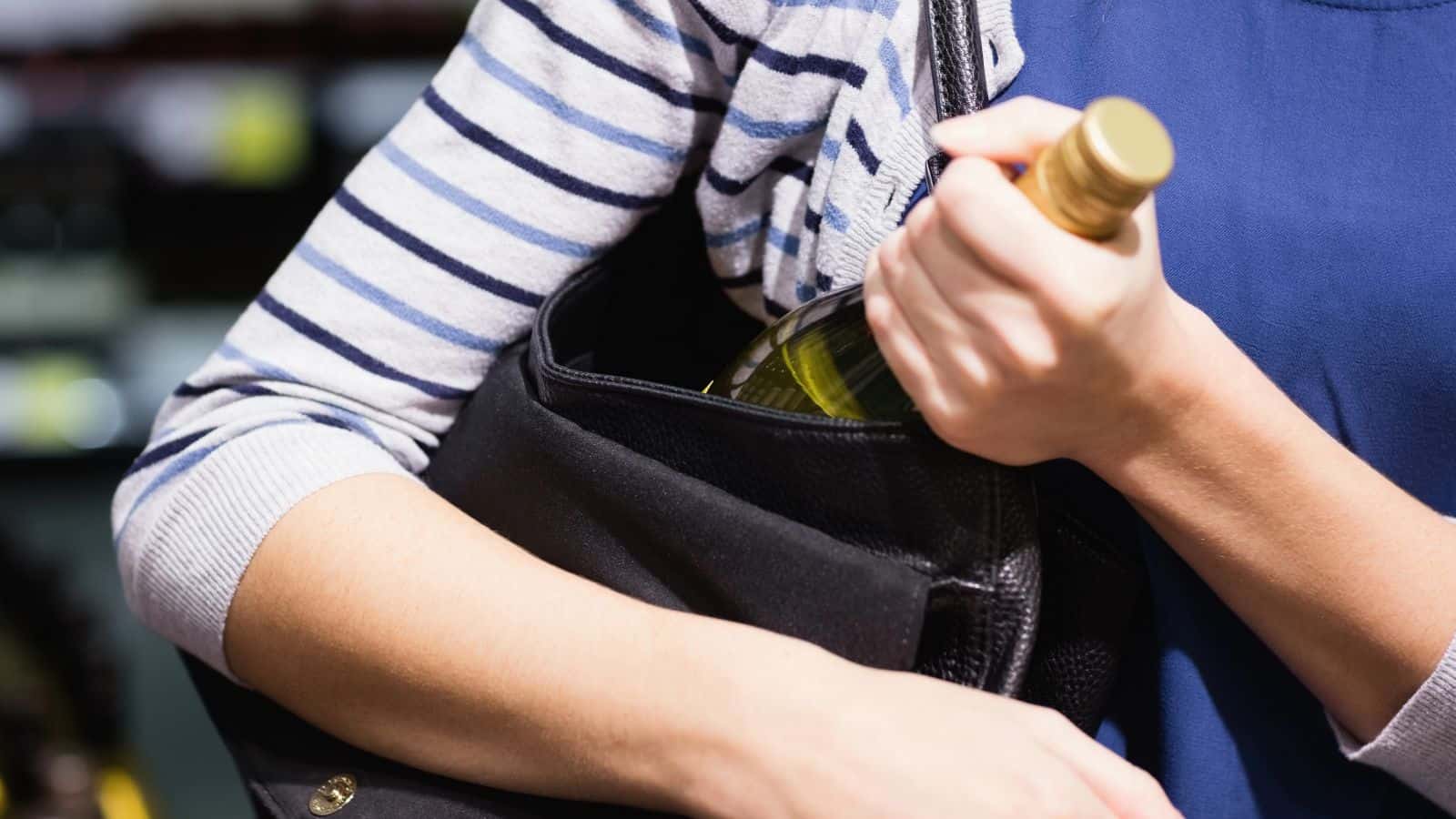 A person wearing a striped shirt is placing a bottle with a golden cap into a black bag. The scene appears to be in a retail setting with blurred shelves in the background.