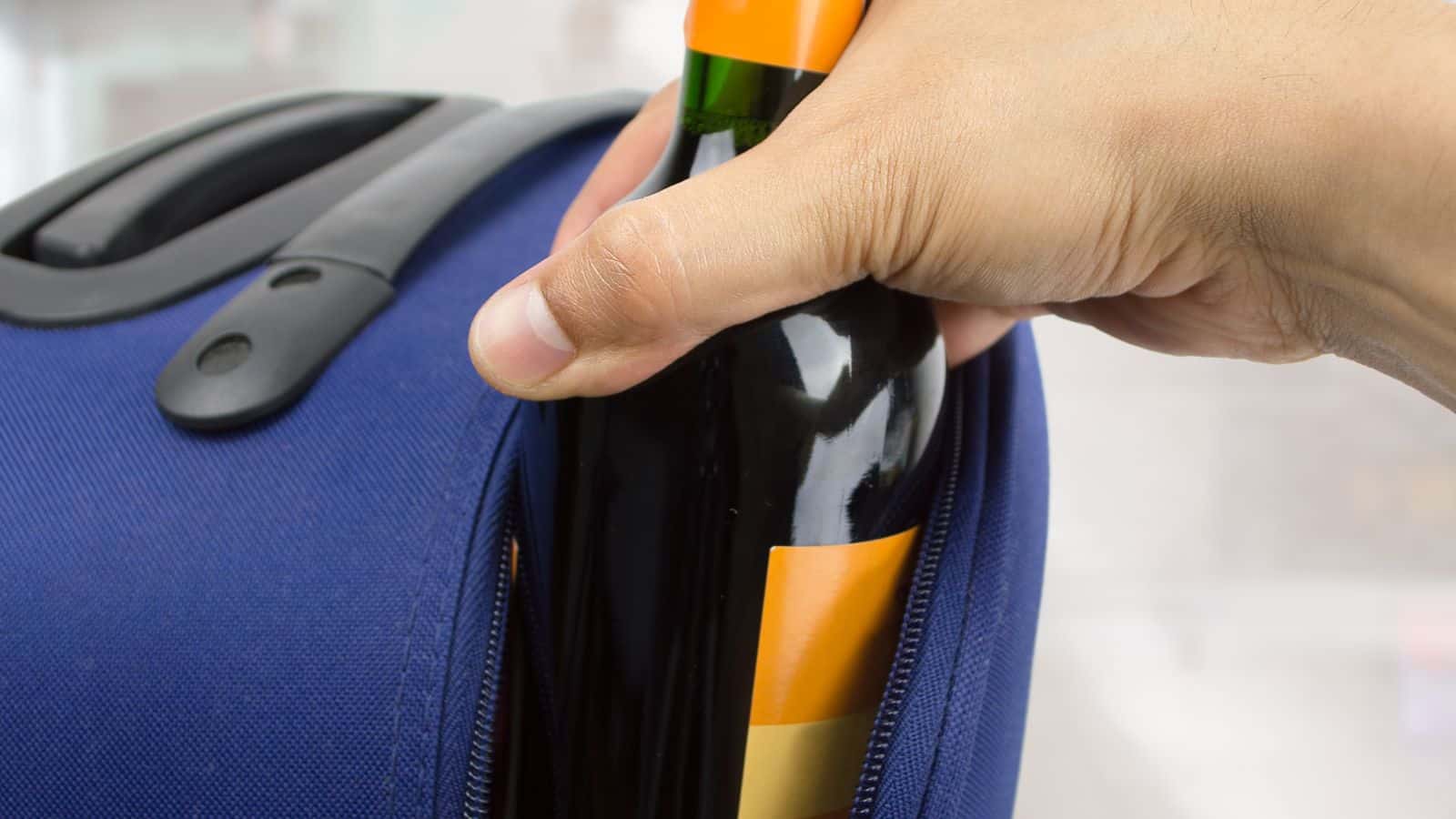 A hand is placing a bottle with a yellow label into the side pocket of a blue backpack. The bottle appears to be made of dark glass, possibly a wine bottle. The backpack is zipped open slightly to fit the bottle.