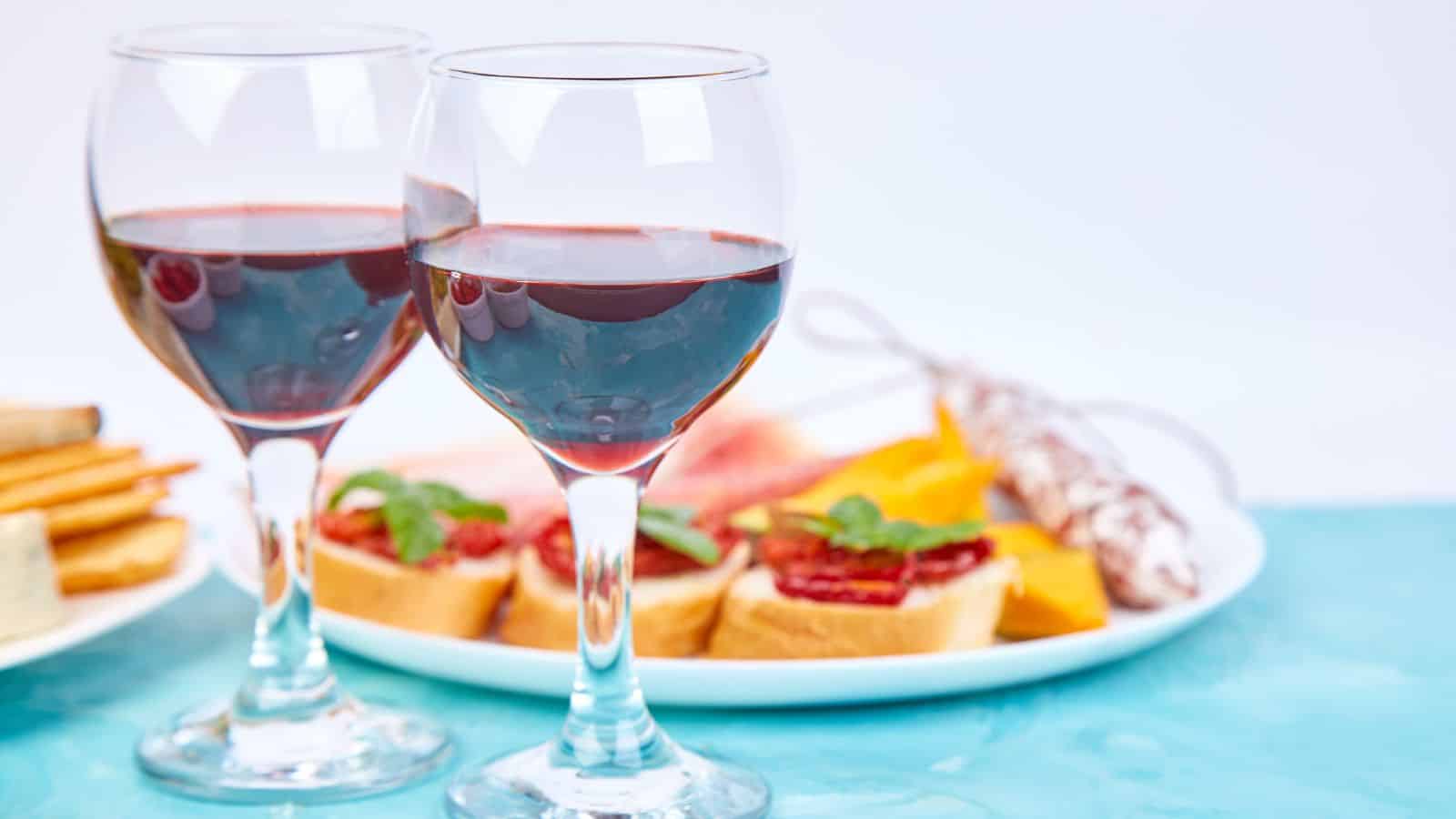 Two glasses of red wine in the foreground. In the background, there are slices of baguette topped with tomato and herbs, alongside pieces of watermelon, yellow fruit slices, and a sausage. The table surface is turquoise.