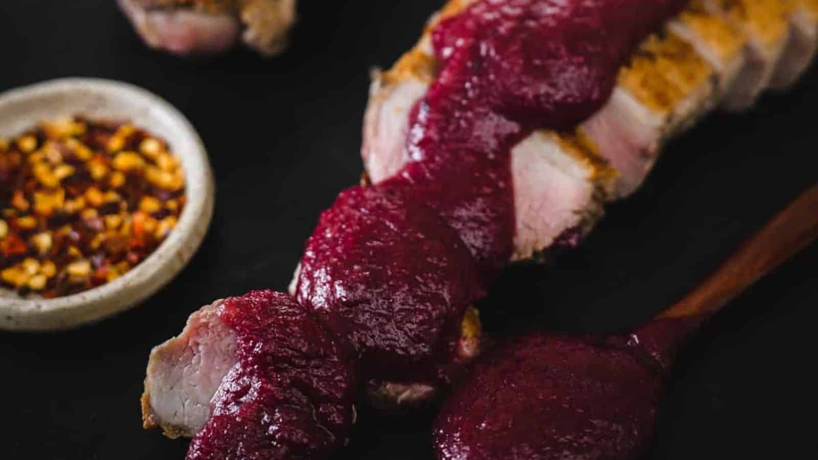 Slices of grilled meat topped with a thick, dark red sauce are arranged on a dark surface. A small bowl of crushed red pepper flakes is beside them. A wooden spoon coated with the sauce rests nearby.