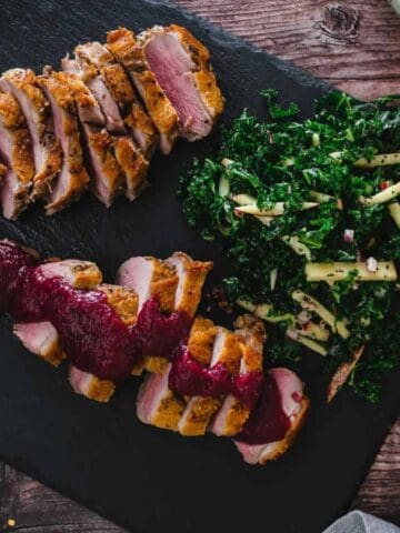 Sliced duck breast with a dark sauce is served on a black slate. Accompanied by a side of kale salad and a small bowl of red pepper flakes. The wooden table background adds a rustic touch.