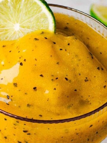 A glass is filled with a thick yellow smoothie, garnished with a lime slice. Another similar glass is partially visible. A halved passion fruit and a lime wedge are in the background on a marble surface.