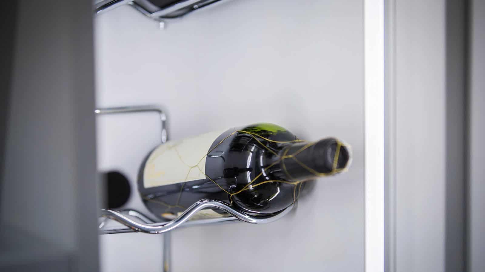 A wine bottle is laid horizontally in a metal rack inside a refrigerator. The bottle is dark with a light label and gold accents. The background is a light, neutral color.