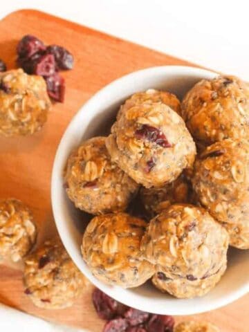 A wooden board holds a white bowl filled with round oatmeal energy bites containing visible oats and cranberries. Additional energy bites, a few oats, and dried cranberries are scattered around the cutting board.