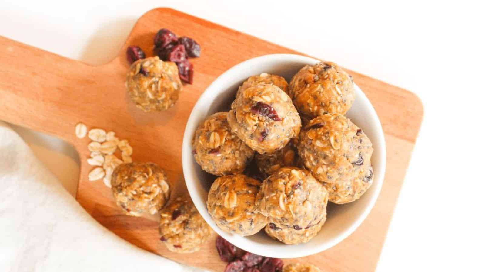 A wooden board holds a white bowl filled with round oatmeal energy bites containing visible oats and cranberries. Additional energy bites, a few oats, and dried cranberries are scattered around the cutting board.