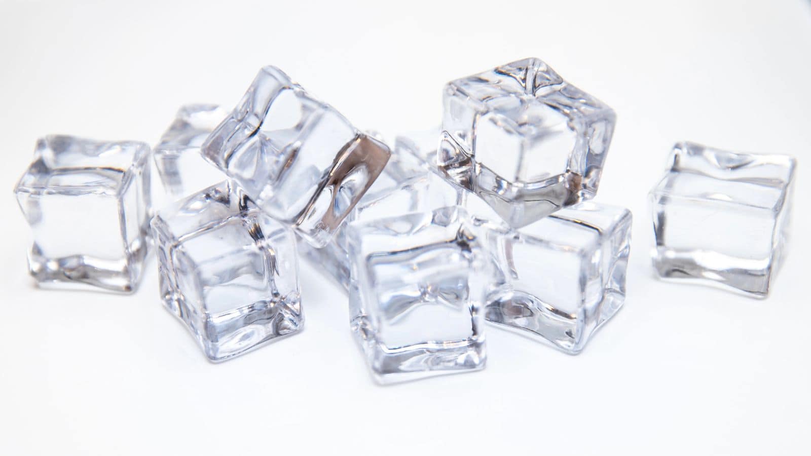 A group of transparent ice cubes piled on a white surface, displaying reflections on their smooth surfaces and slightly rounded edges.