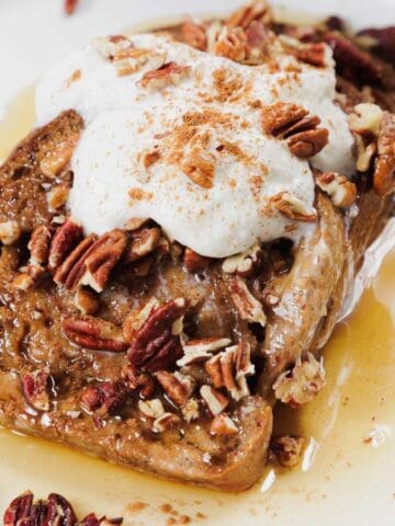 A slice of French toast topped with whipped cream, chopped pecans, and a sprinkle of cinnamon, drizzled with syrup on a white plate.