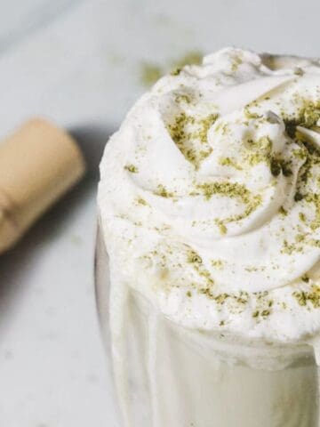 A glass filled with a creamy matcha drink topped with swirls of whipped cream and sprinkled with matcha powder. A paper straw is inserted into the drink. A bamboo whisk with matcha powder around it is in the background.