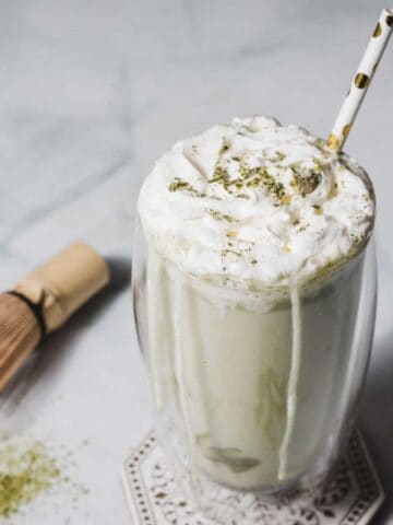 A glass of matcha latte topped with whipped cream and matcha powder sits on a table. A spotted straw is inserted into the drink. A bamboo whisk and a pile of matcha powder are nearby on the table.