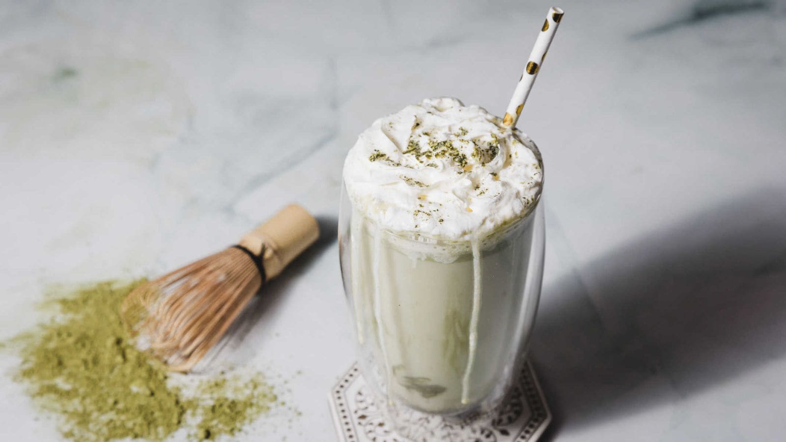 A glass of matcha latte topped with whipped cream and matcha powder sits on a table. A spotted straw is inserted into the drink. A bamboo whisk and a pile of matcha powder are nearby on the table.