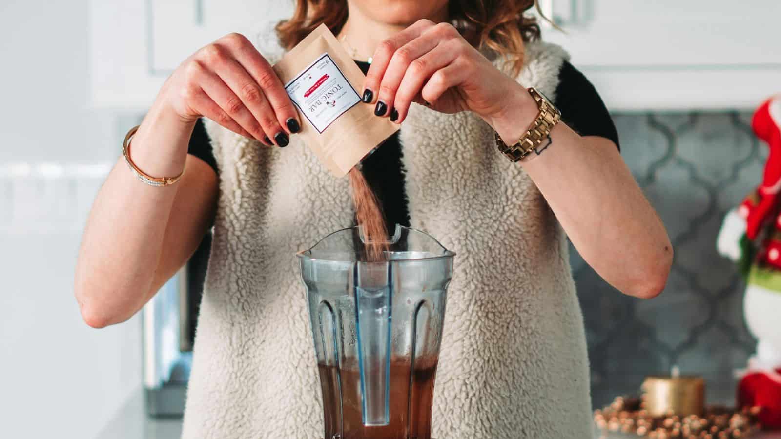 A person wearing a fuzzy beige vest is pouring a packet of chocolate powder into a blender filled with liquid. The kitchen counter has a festive decoration in the background, and the person has black painted nails and wears a watch and bracelets.