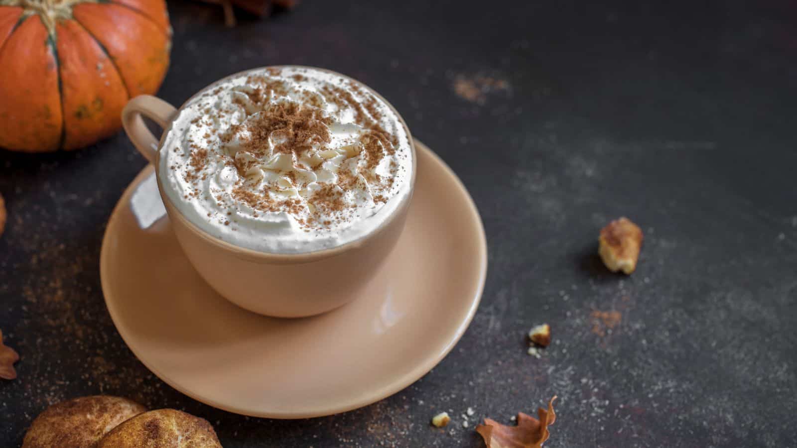 A beige cup filled with whipped cream-topped coffee or latte. The drink is sprinkled with cinnamon. It sits on a matching saucer on a dark surface. A small pumpkin and scattered autumn leaves are nearby.