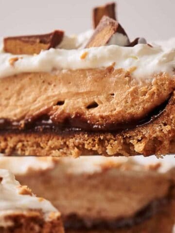A slice of a layered dessert is being lifted from the whole. It has a crumbly crust, a chocolate layer, a thick peanut butter filling, and a topping of whipped cream. Chocolate pieces are scattered on top.