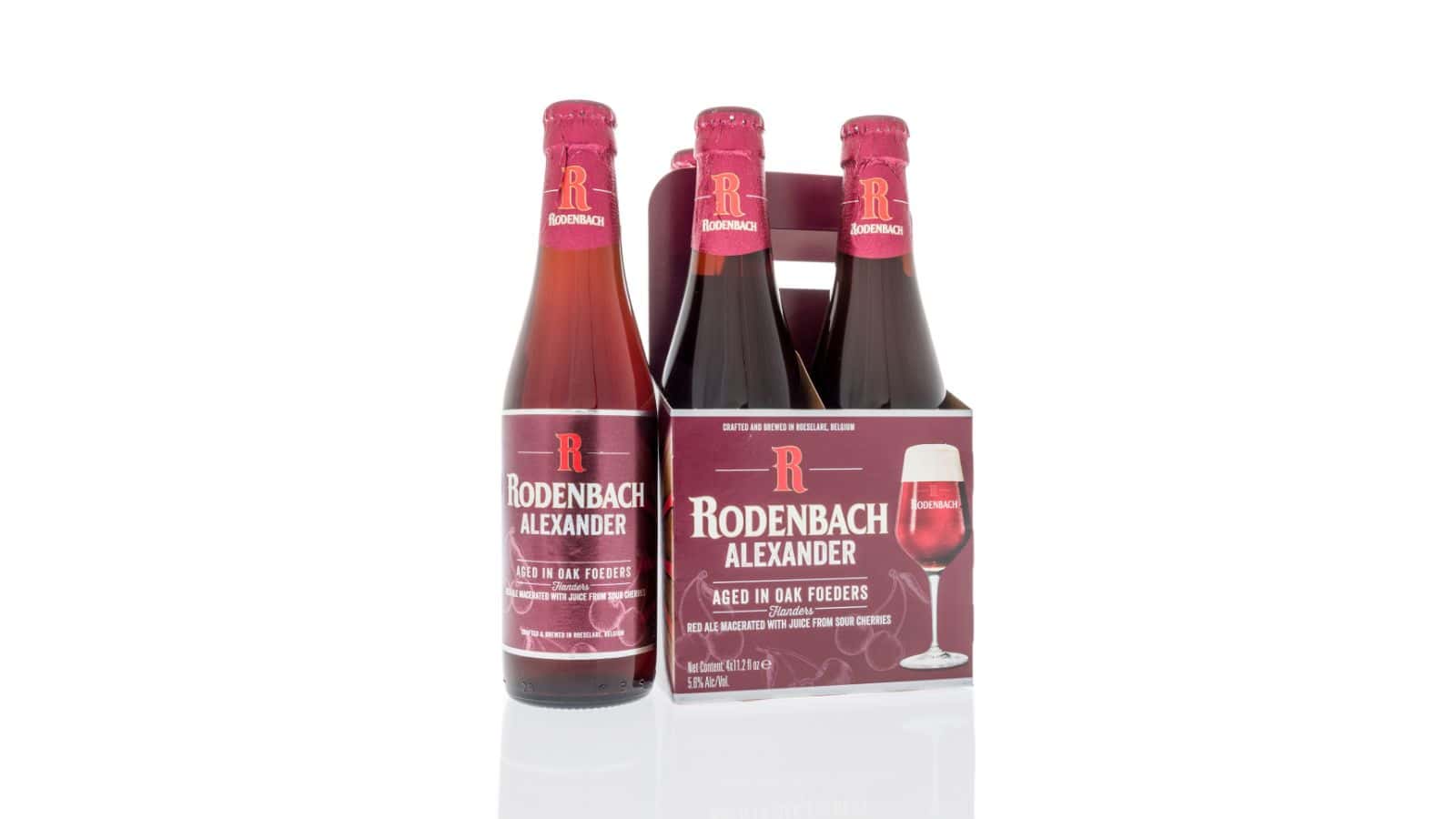 A four-pack of Rodenbach Alexander beer bottles is shown. The bottles have red caps and labels with white text. They are held in a matching cardboard carrier with an image of a beer glass on the side. The background is white.