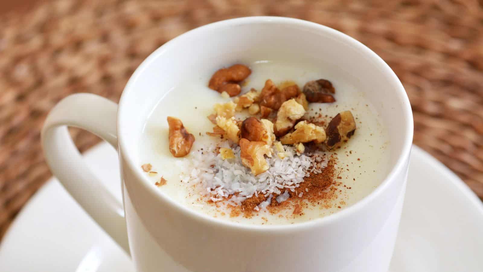 A white mug filled with creamy rice pudding, topped with chopped nuts, shredded coconut, and a sprinkle of cinnamon. The mug sits on a white saucer, with a woven texture background.