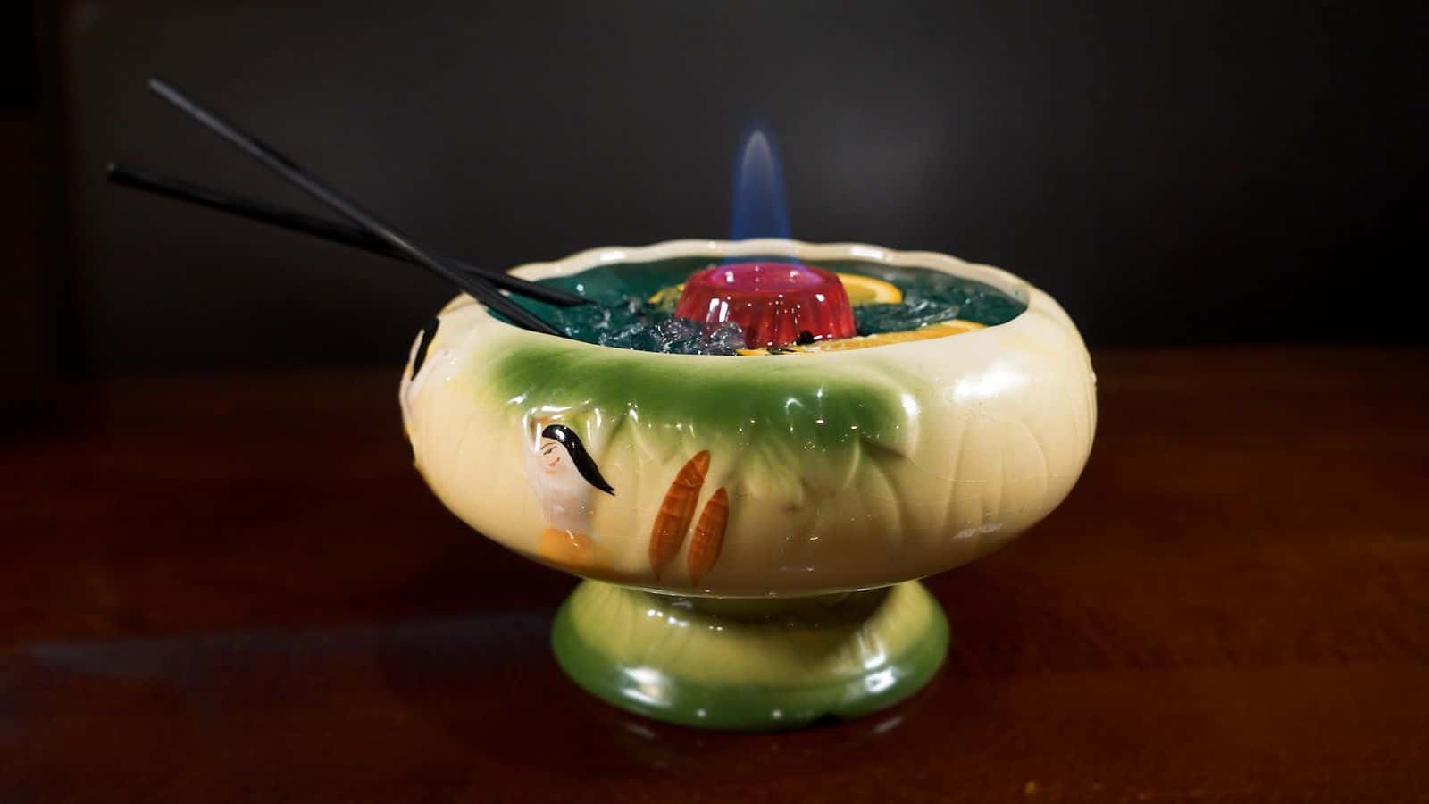 A decorative ceramic bowl with a tropical design holds a mixed drink with a red center and two black straws. The drink is on fire, and there are visible pieces of fruit and crushed ice.