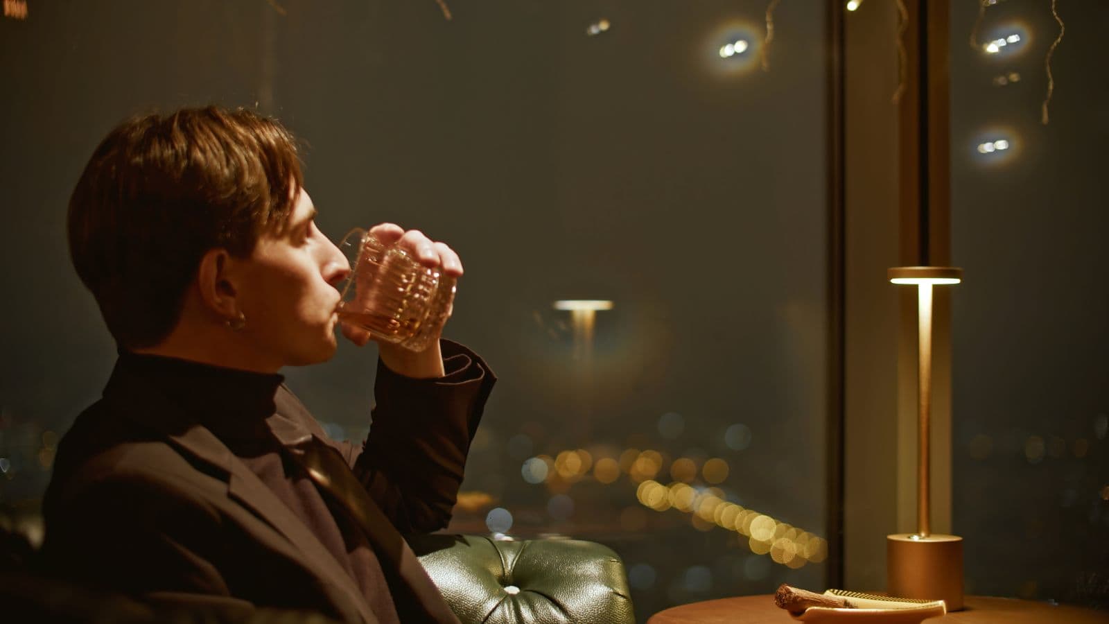A person in a dark suit sits on a leather chair, drinking from a glass in a dimly lit room. A window reveals city lights in the background. A small lamp and an item placed on a wooden table are visible beside the person.