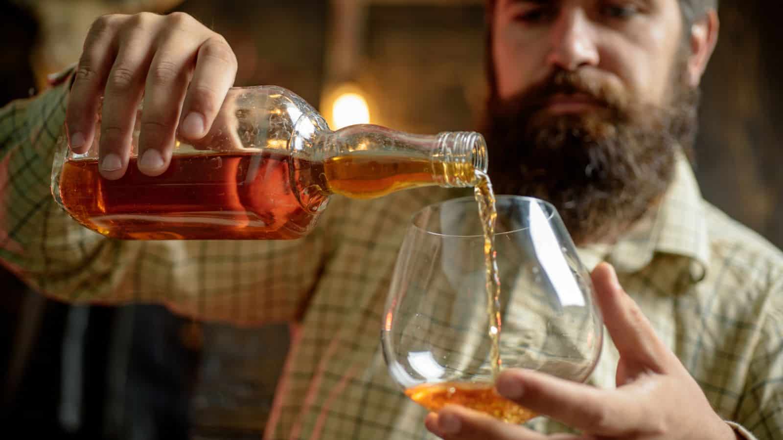 A man with a beard pours a dark amber liquid from a bottle into a large glass. He is wearing a plaid shirt and is focused on his task. The background is softly blurred with warm lighting.