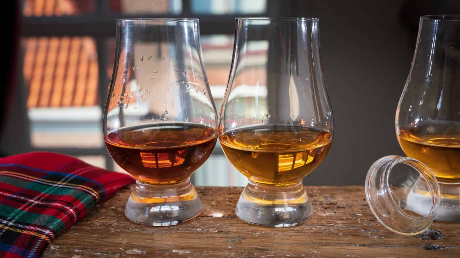 Two glasses of whiskey are placed on a wooden table, each filled with a different amber-colored liquid. A clear lid is beside the second glass. A plaid cloth is to the left, and a blurred window background shows buildings.