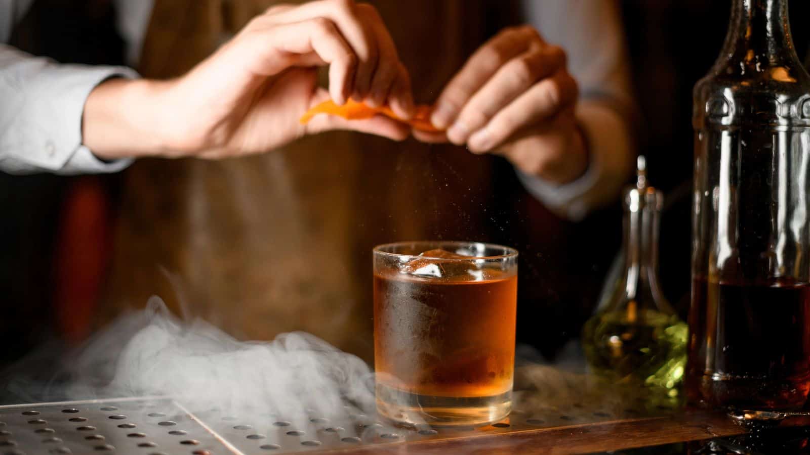 A person in an apron prepares a cocktail by squeezing an orange peel over a glass with ice. The drink is surrounded by mist, and there are bottles of alcohol nearby on the bar counter.