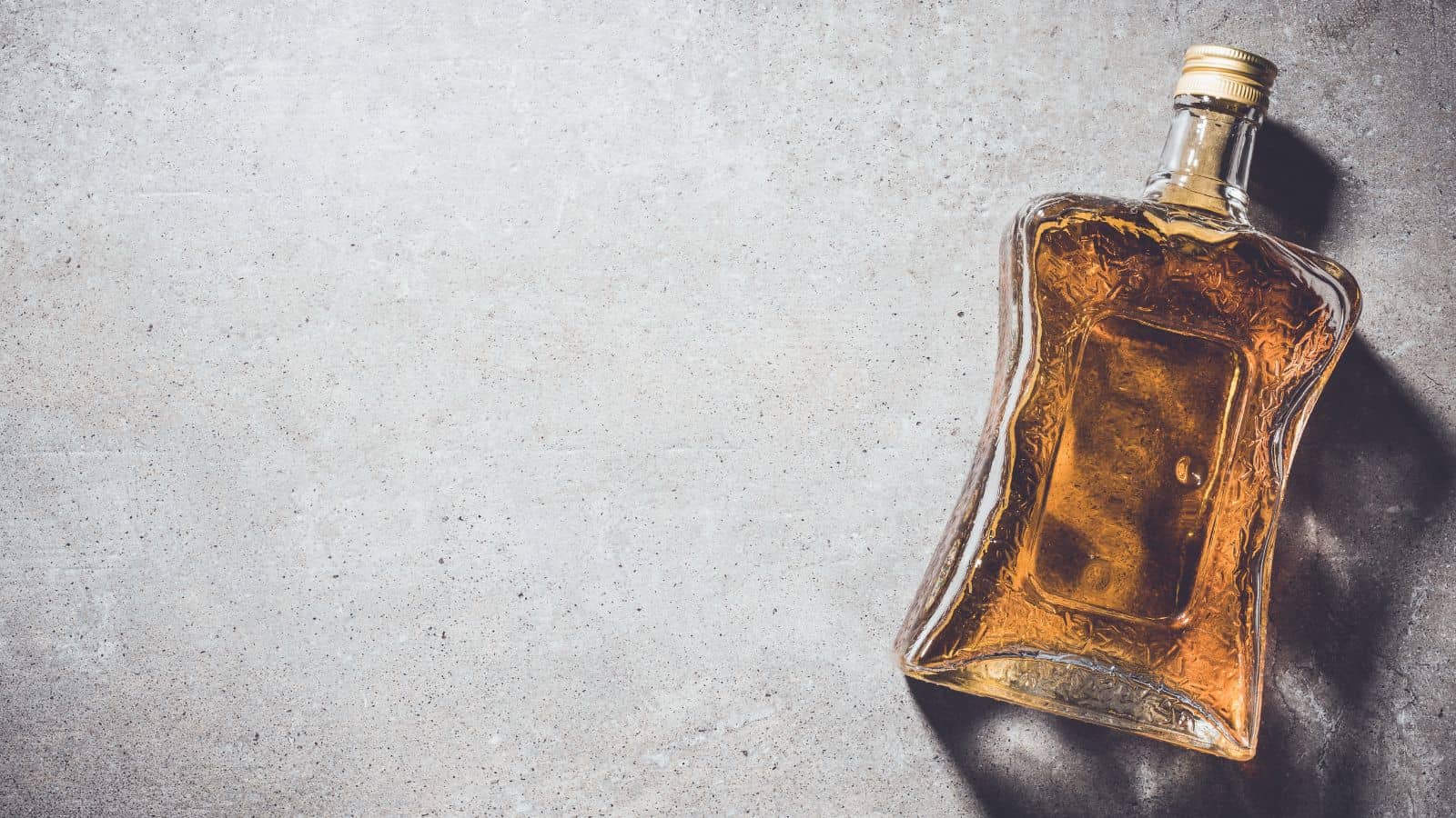 A glass bottle filled with amber liquid is lying on a gray textured surface. The bottle has a rectangular shape with curved edges and a gold cap. Shadows from the bottle fall to the right. The image has a minimalist composition.