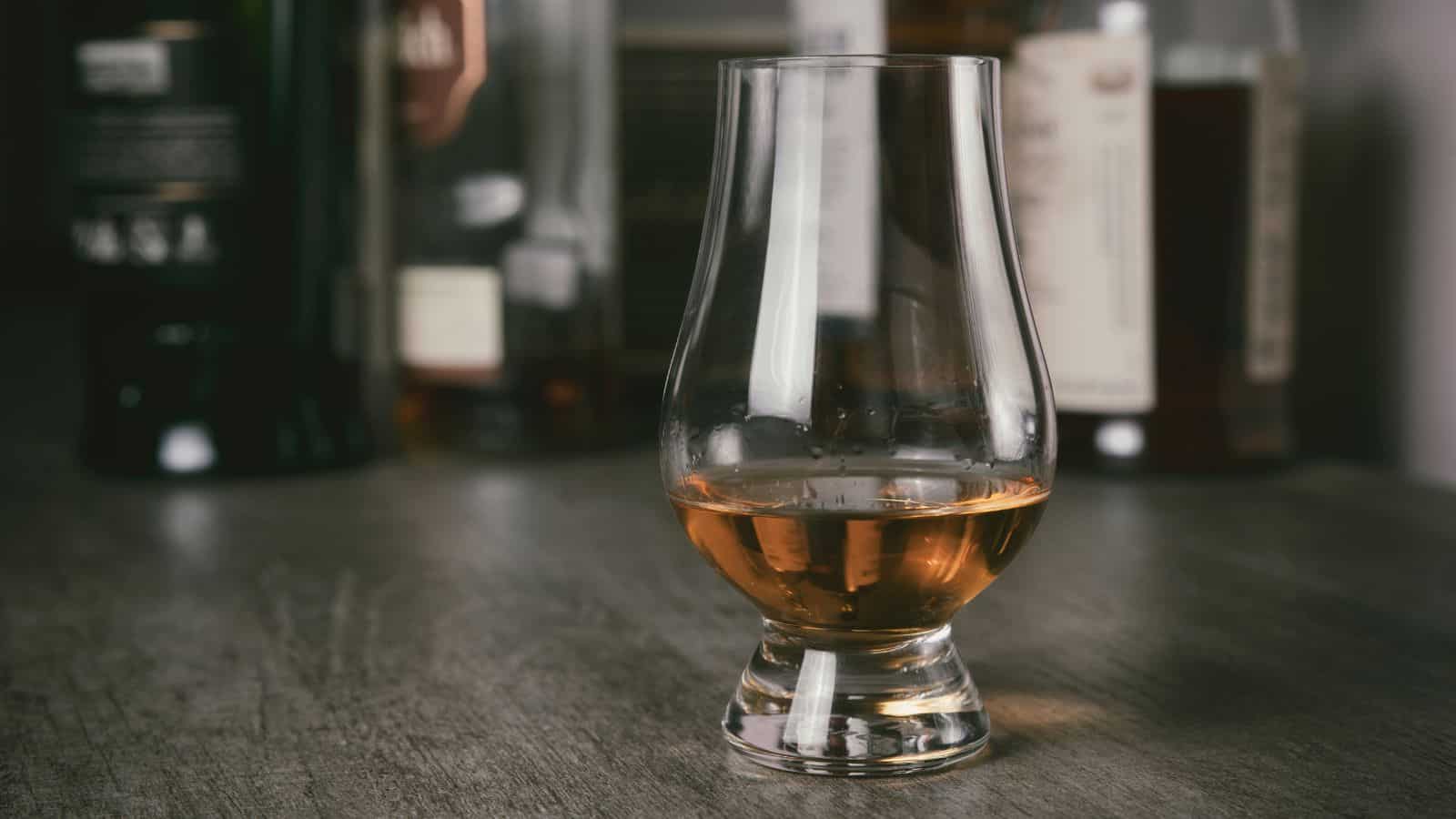 A clear tulip-shaped glass with a short stem contains amber liquid, likely whiskey, on a wooden surface. Several blurred bottles in the background suggest a bar or tasting setting.