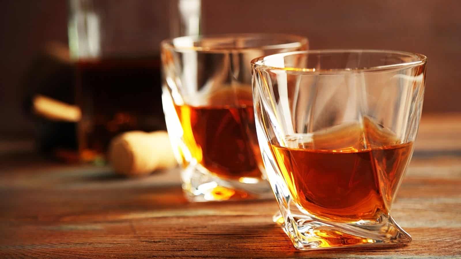 Two glasses of amber-colored whiskey are placed on a wooden surface. A cork and a partially visible bottle are in the background. The glasses have a distinctive square shape with twisted sides.