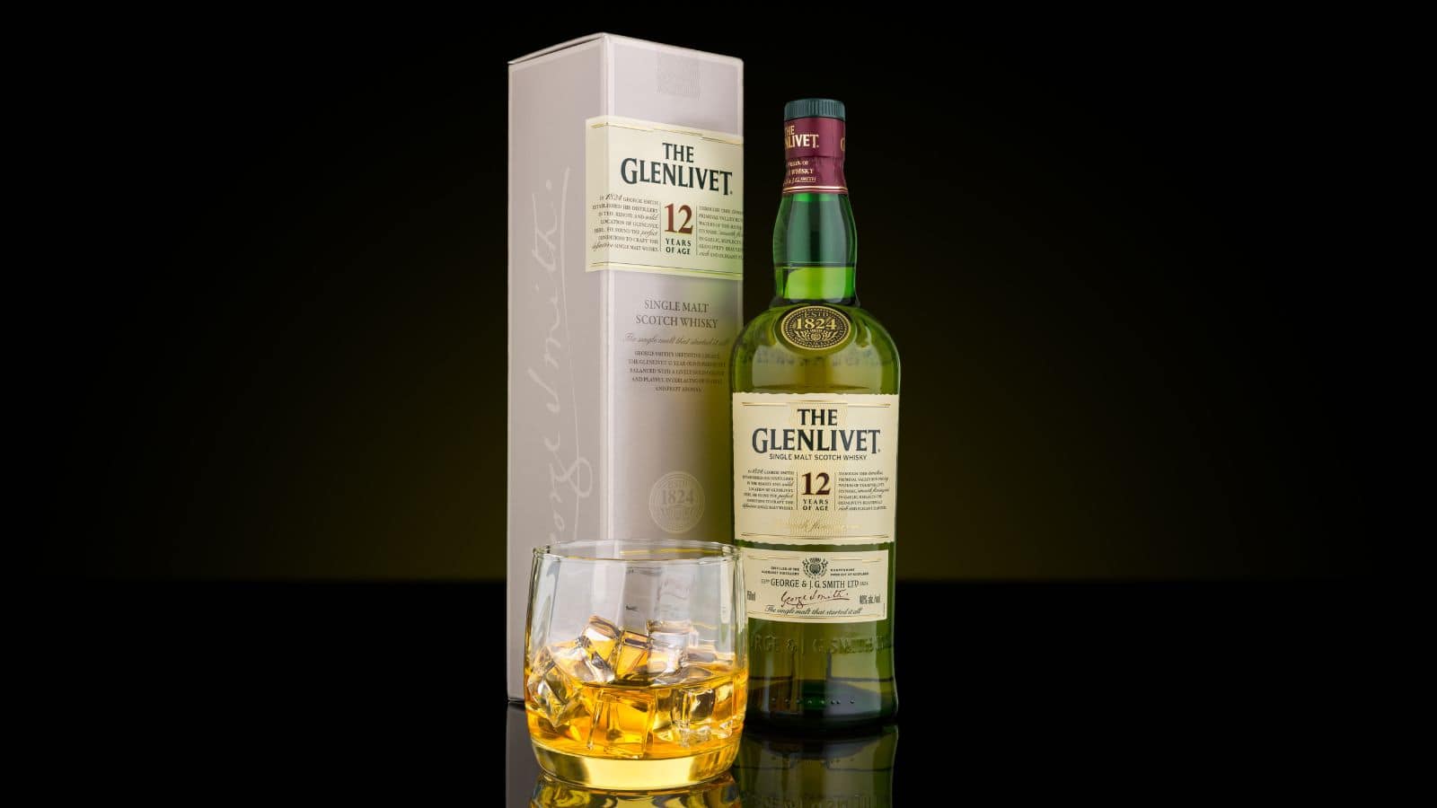 A bottle of The Glenlivet 12 Year Old Single Malt Scotch Whisky is displayed next to its box. In front, a glass filled with the whisky and ice cubes is placed. The background is dark, highlighting the items.