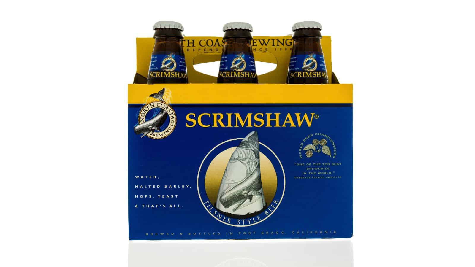 A six-pack of Scrimshaw Pilsner Style Beer in a blue and yellow cardboard carrier. The packaging features an image of a carved whale tooth. The bottles are from North Coast Brewing Company, labeled as having water, malted barley, hops, and yeast.