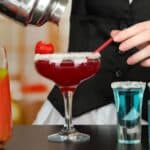 A person wearing a black vest pours a red drink from a shaker into a margarita glass with a sugared rim and a strawberry garnish. Nearby are three blue drinks in glassware and a tall glass of red beverage with a lime slice and straw.