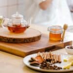 A glass teapot filled with tea sits on a wooden board next to a jar of honey with a dipper. In the foreground, a plate holds cinnamon sticks, lemon wedges, and dried fruit slices. A person is blurred in the background.