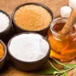 Various sweeteners are displayed on a wooden surface. There are bowls containing white sugar, brown sugar, honey with a dipper, a white powdered substance, and orange granules. Sugar cubes and green leaves are also present.