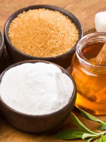 Various sweeteners are displayed on a wooden surface. There are bowls containing white sugar, brown sugar, honey with a dipper, a white powdered substance, and orange granules. Sugar cubes and green leaves are also present.