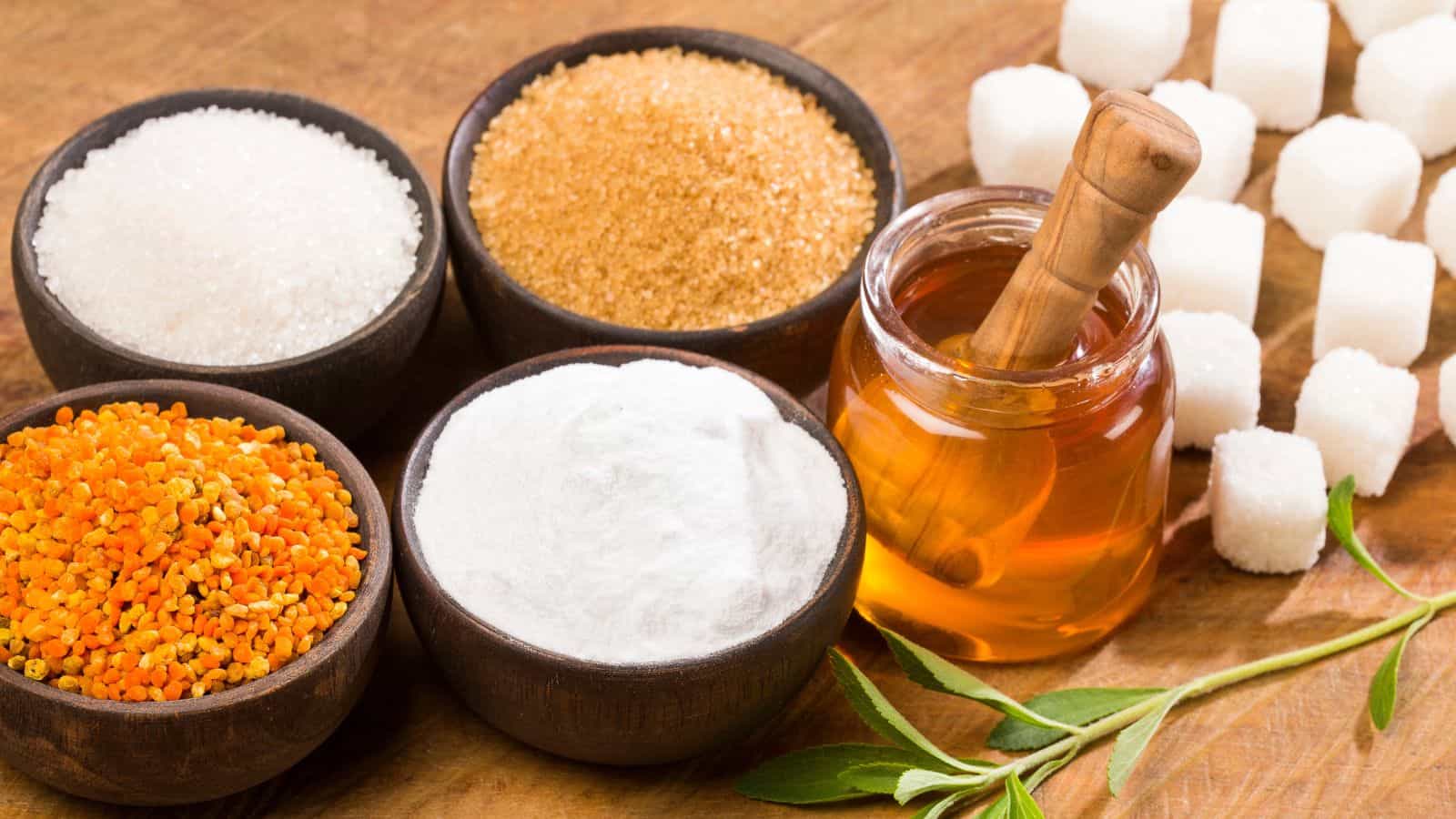 Various sweeteners are displayed on a wooden surface. There are bowls containing white sugar, brown sugar, honey with a dipper, a white powdered substance, and orange granules. Sugar cubes and green leaves are also present.