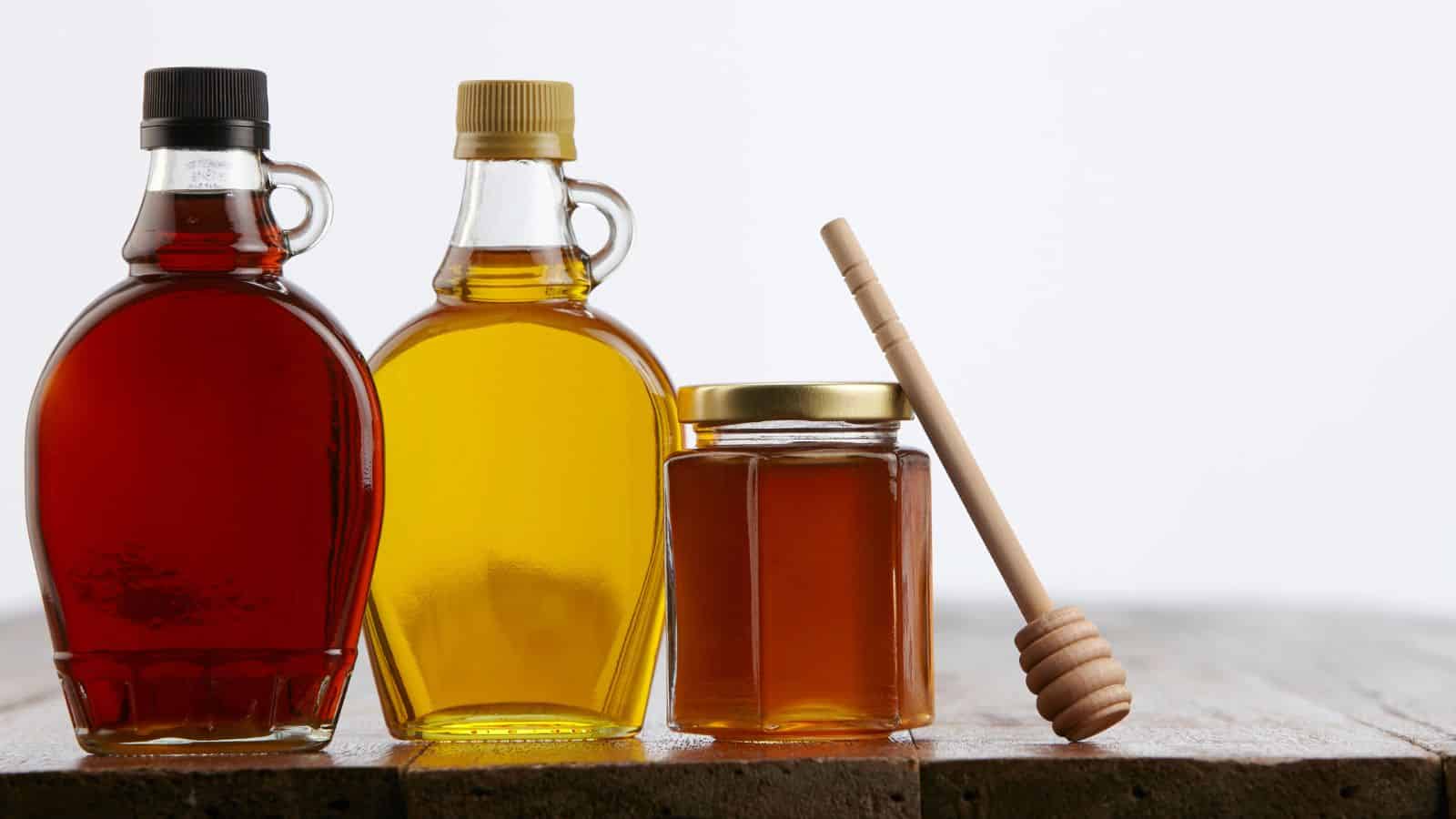 Two glass bottles and a jar filled with amber liquids are on a wooden surface. The first bottle has a darker liquid and a looped handle, the second has a lighter hue with a yellow cap. A wooden honey dipper rests beside the jar.